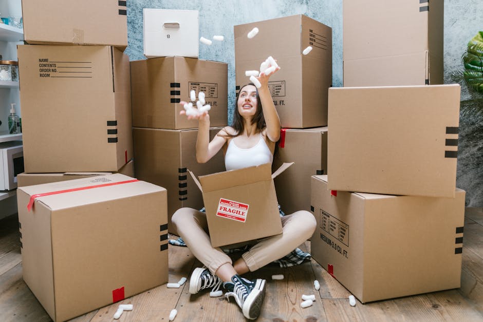 A young woman with long dark hair, dressed in a white sleeveless top and beige pants, is sitting cross-legged on a wooden floor inside a room surrounded by numerous brown cardboard boxes of various sizes, some labeled with 'fragile' stickers. She is inside an open box and is actively throwing white packing peanuts into the air, with some falling around her. The boxes are stacked on the floor and also against the wall, which has a textured, light-colored finish. A green potted plant is partially visible on the right side of the image, and there is a small white counter or shelf on the left with decorative items. This scene captures the packing process in preparation for house removals, highlighting the content handling and整理 involved in home relocation, as managed by Harringay Man and Van for efficient furniture transport and move logistics.