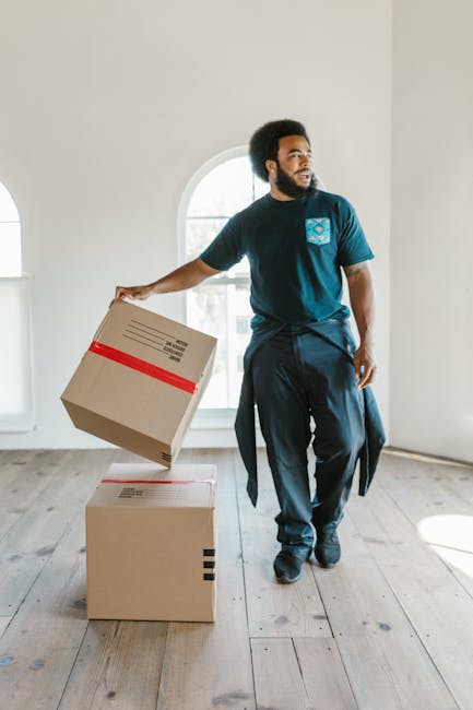 A man with curly hair, wearing a blue t-shirt and dark trousers, is sitting on a wooden floor inside a room next to a large potted plant. He is surrounded by several cardboard boxes, some sealed with red packing tape, which are stacked and placed near the window. The room has high, arched windows allowing natural light to illuminate the space, with a view of parked cars and part of a building outside. The scene reflects a moving or packing process, typical of home relocation or furniture transport preparations, as facilitated by Harringay Man and Van. The wooden floor and minimal interior detail emphasize the focus on the packing activity and the transition phase in a house removal service.