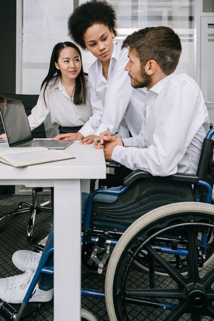 An indoor office setting featuring two women and one man with a disability. The man, seated in a wheelchair, is engaged in a conversation with a woman standing next to him, who is leaning slightly forward and making eye contact. The woman sitting across the table has long black hair and is attentively observing the interaction. The table has a laptop, an open folder, and some documents on it. The background reveals large windows with blinds, allowing natural light to illuminate the space. This scene may depict a discussion related to work, accessibility, or support services, reflecting inclusive professional environments. The setting underscores the importance of accessible workplaces and effective communication, relevant to service providers like Harringay Man and Van involved in home relocation and furniture transport, especially when considering accessibility and moving logistics.