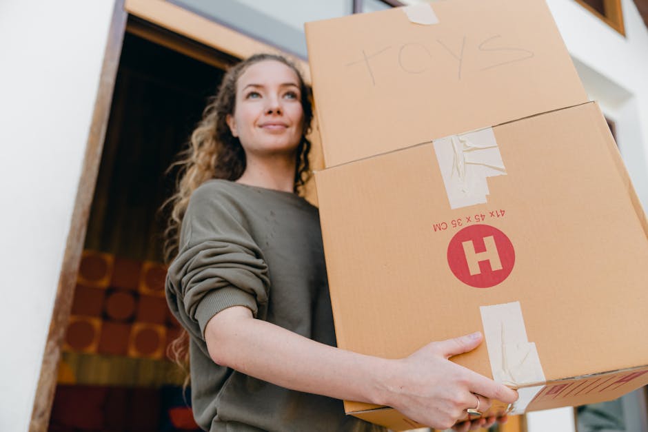 A woman with curly brown hair wearing a grey long-sleeved top is standing outside a building, holding a large cardboard box labeled 'toys' with masking tape. Behind her, there is a dark door frame and the interior of the property is partially visible. She is preparing for a home relocation or moving process, with the box indicating packing for moving services. The scene is lit with natural daylight, highlighting the details of the box, her facial expression, and surroundings. The image emphasizes the early stages of packing and the logistical aspects of furniture transport and house removals, consistent with the services offered by Harringay Man and Van as seen on their website.