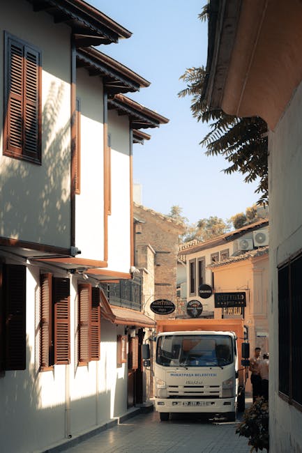 A narrow street scene outside a residential building during daytime, with a white moving van positioned in the middle of the alleyway, partly blocking the view of the building's entrance. The building has a white facade with brown window shutters and a sloped roof with overhanging eaves. To the right, another building with a similar white exterior and a small window is visible, along with a person standing near the van, possibly supervising or assisting with the move. The street is paved with light-colored bricks, and nearby signs indicate local businesses. Trees and additional buildings are seen in the background under a clear sky. Harringay Man and Van, providing home relocation and furniture transport services, are involved in the loading process, arranging boxes and furniture for transport, typical of house removals with narrow access. The scene captures the logistics of packing, lifting, and loading household items in a compact, built-up urban environment.
