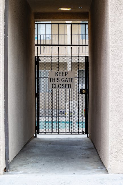 A narrow outdoor passageway with beige stucco walls leading to a metal gate secured with a sign that reads 'Keep This Gate Closed'. Beyond the gate, an apartment or condominium complex with a balcony and windows is visible. The ground is concrete, and there is no visible debris or obstruction. The scene is well-lit with natural daylight, and the gate appears to be part of a property boundary or communal entrance. This image illustrates a typical entry point in a residential building that Harringay Man and Van might encounter during home relocation or furniture transport tasks involving buildings with limited access.