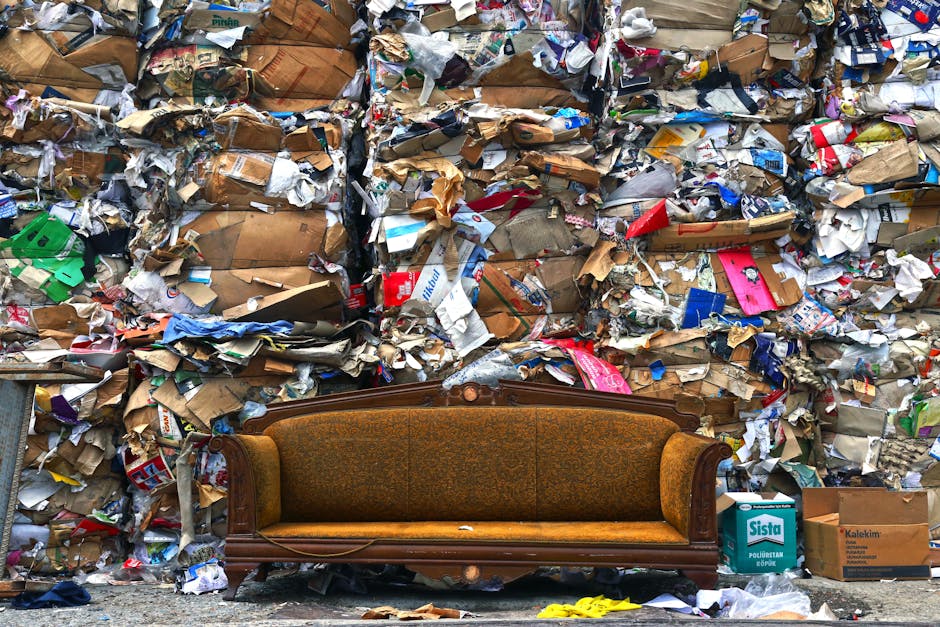 An aerial view of a large pile of mixed household waste and debris on the ground, consisting of crumpled cardboard boxes, plastic containers, paper, and discarded packaging materials, spread across a
