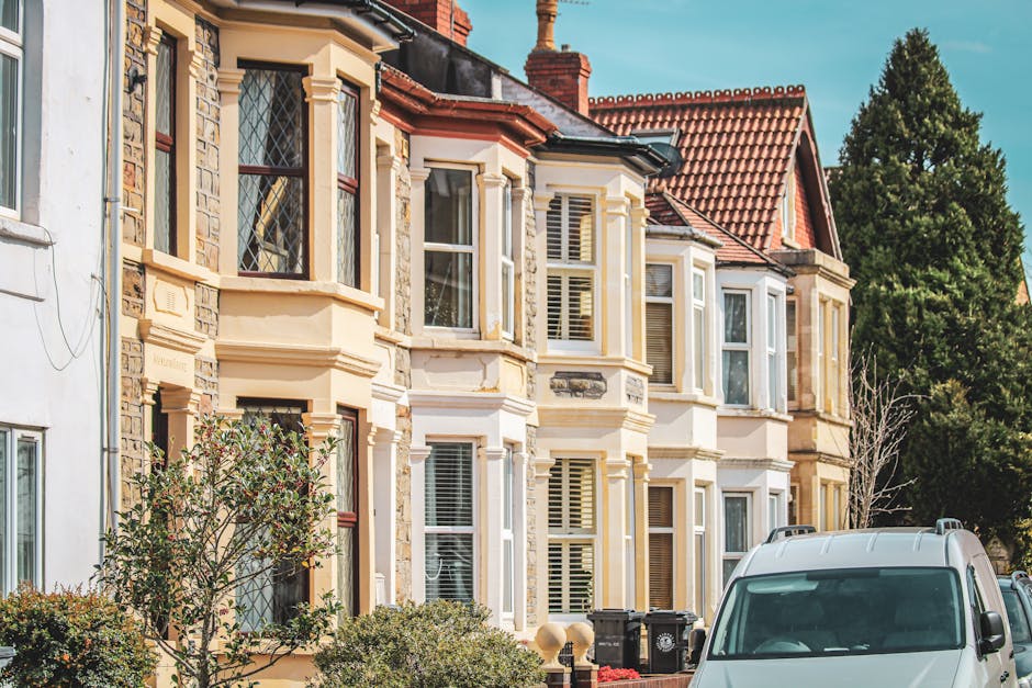 A row of Victorian terraced houses with ornate bay windows and detailed stonework on a residential street, showcasing a typical entrance with front gardens and greenery, under a clear blue sky. In the foreground, part of a white van is visible, likely used by Harringay Man and Van during a home relocation or furniture transport service. The scene suggests a house removal process, with the van parked close to the property, ready for loading or unloading. The image captures the exterior environment including nearby trees and pavement, emphasizing the urban setting for professional moving and packing activities.