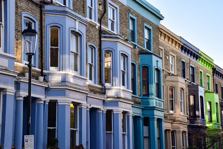 A row of Victorian terraced houses with bay windows and ornate architectural details, painted in various pastel shades including light blue, beige, and green, located on a residential street. Outside, a black metal lamppost stands on the pavement. The windows reflect the sky and nearby buildings, with some showing curtains or blinds. In front of one house, there is a partially visible driveway or small area for parking, with a parking permit sign attached to a lamppost. As part of the home relocation process, Harringay Man and Van is seen preparing for furniture transport, with the possibility of loading or unloading belongings from the street or driveway area. The setting includes clear natural lighting, emphasizing the different colours and architectural features of the houses, indicative of a typical London neighbourhood preparing for or engaged in a house removal or moving service involving packing, loading, and furniture transport within the property or onto a removal vehicle.
