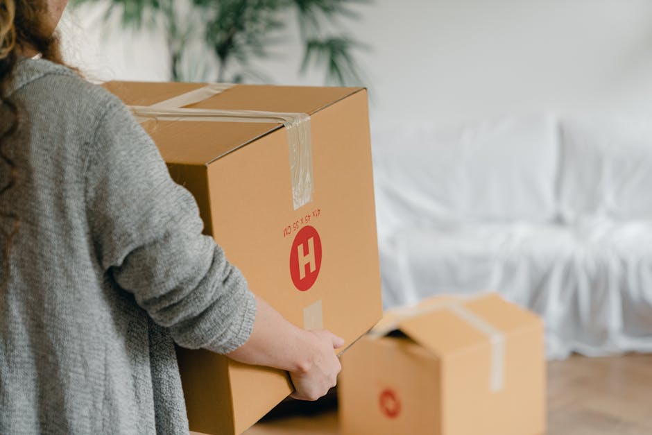 Close-up of a person holding a medium-sized cardboard box sealed with clear packing tape, with a large red ‘H’ logo and some printed text visible on the side, inside a room with a wooden floor and a white fabric-covered sofa in the background. Additional smaller boxes are seen on the floor nearby, indicating packing and home relocation activities. The person is wearing a grey knitted sweater, and the scene suggests the process of loading or unpacking during furniture transport or house removals, exemplifying procedures involved in professional removals services such as those offered by Harringay Man and Van, supporting efficient packing and moving logistics.