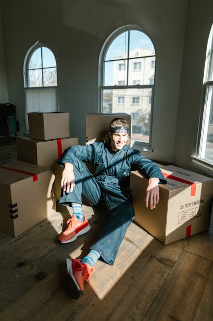 A young man wearing a blue work uniform, a patterned headband, and bright orange sneakers sits on a wooden floor inside a well-lit room with large windows. Surrounding him are several large cardboard boxes sealed with red and black packing tape, indicating an ongoing home relocation process. The boxes are stacked and arranged around him, suggesting they are ready for transport or unpacking as part of furniture transport and packing and moving activities. The sunlight streaming through the windows creates a natural, warm atmosphere, highlighting the movement and preparation involved in a house removal. This scene reflects the logistical effort of loading, packing, and organizing belongings during a professional move facilitated by Harringay Man and Van, a company specialising in removals and relocation services in the area.