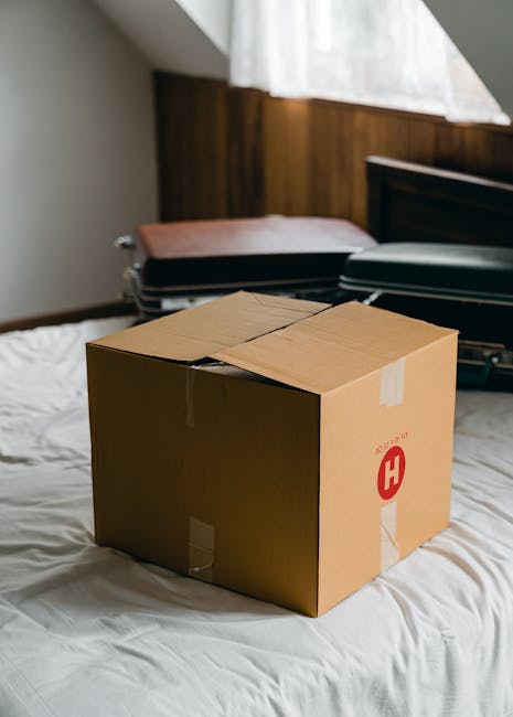 A cardboard moving box sealed with packing tape sits on a white bedspread in a bedroom with wooden wall paneling and natural light entering through a window with sheer curtains. Behind the box, there are two closed suitcases and an open black suitcase with visible interior compartments, indicating packing and home relocation activity. The scene suggests a furniture transport or packing process as part of an overall moving service, likely handled by Harringay Man and Van, specializing in same-day flat moves and urgent house removals in Harringay. The environment appears temporary and prepared for loading onto a van, with a focus on the logistical steps involved in the packing and moving process.