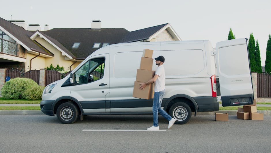 A man wearing a white T-shirt, blue jeans, and a black cap is seen outside a white cargo van, which is parked on a residential street in front of suburban houses with pitched roofs and dormer windows. The man is carrying three stacked cardboard boxes, supporting them with both hands as he walks away from the open side door of the van. Several additional cardboard boxes sit on the pavement near the van’s rear and side, suggesting the process of home relocation or packing and moving. The van’s side door is open, indicating the loading process, and the surroundings include well-maintained lawns, a fenced garden, and tall green trees. The lighting is natural, typical of daylight, and the scene depicts a typical house removals operation, with a focus on furniture transport and packing logistics, supported by Harringay Man and Van’s professional removals service.