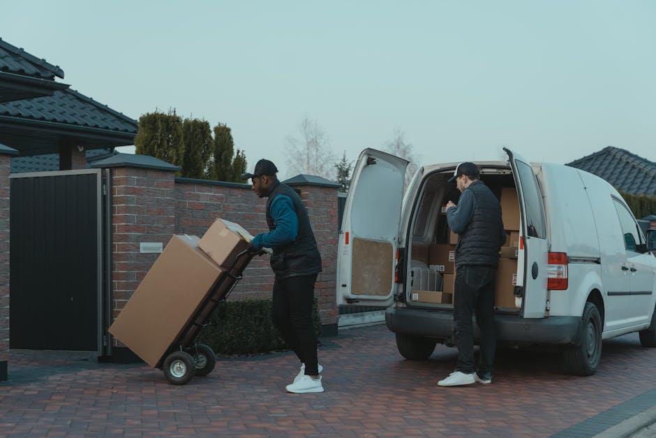 Two movers from Harringay Man and Van are engaged in a home relocation process outside a residential property during daytime. One individual, wearing a black cap and a blue and black jacket, is pushing a two-wheeled trolley loaded with a large cardboard box towards the driveway entrance, which is paved with red bricks. The second person, dressed in a black cap and jacket, is standing beside an open white van, which is parked on the driveway with its rear doors wide open, revealing several packed cardboard boxes inside. The scene is set in a suburban neighbourhood with houses featuring dark tiled roofs and brick or wooden fencing. The area appears to be well-lit with natural daylight, and the environment indicates a typical house removal operation involving the careful loading and transport of household items, supported by packing materials like cardboard boxes, which are visible inside the van. Harringay Man and Van is actively involved in furniture transport and packing and moving logistics during this staged loading process.