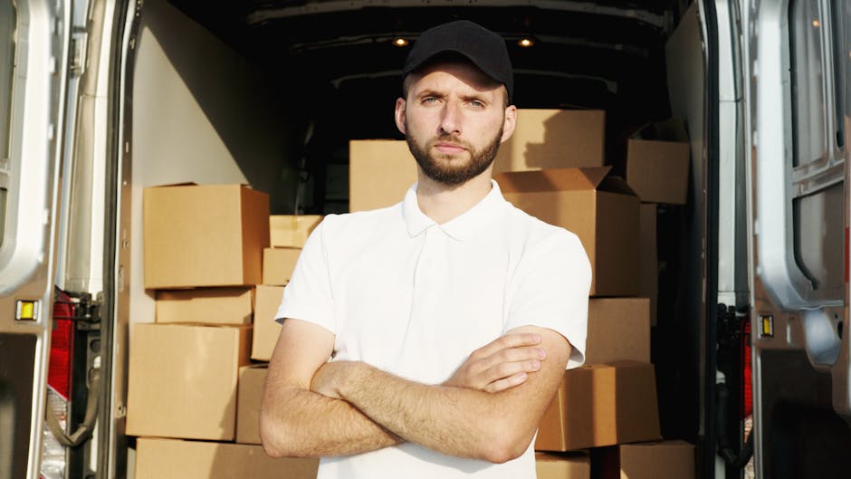 A young man with a serious expression, wearing a black cap and a white polo shirt, stands with his arms crossed inside the open back of a delivery van loaded with numerous cardboard moving boxes of various sizes, some with packing tape, and plastic wrapping. The van's interior is dimly lit with additional boxes stacked further inside, suggesting an ongoing packing or furniture transport process as part of a house relocation. The scene is set outdoors on a pavement, with sunlight illuminating the boxes and the man's face, indicating daylight hours. This image highlights the logistics of packing and loading boxes for a home removal service, providing context for furniture transport and moving activities associated with services offered by Harringay Man and Van, aligned with the broader topic of managing delays in booking removals in Harringay.
