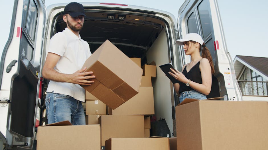 A man and a woman are engaged in a home relocation process, standing at the back of a white moving van with its rear doors open. The man, wearing a white polo shirt, light blue jeans, and a dark baseball cap, is holding a medium-sized cardboard box, positioning it to be loaded or unloaded. Inside the van, there are several other cardboard boxes of various sizes, some partially visible, along with a large white panel and packaging materials such as bubble wrap or plastic sheeting. The woman, dressed in a black sleeveless top, denim shorts, and a white cap, is standing to the right, looking at a tablet device, possibly coordinating the loading process or managing inventory. The van is parked on a driveway or street near a residential area, with a house visible in the background, featuring a tilled roof, window, and partial front yard. Lighting suggests daytime with natural sunlight illuminating the scene. This image is relevant to house removals, packing and moving logistics, and furniture transport services offered by Harringay Man and Van, primarily associated with the page 'What to know about delays with Harringay man and van bookings' on harringaymanandvan.com.