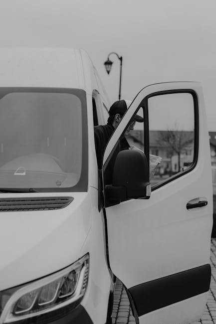 A young man with a serious expression, wearing a black cap and a white polo shirt, stands with his arms crossed inside the open back of a delivery van loaded with numerous cardboard moving boxes of various sizes, some with packing tape, and plastic wrapping. The van's interior is dimly lit with additional boxes stacked further inside, suggesting an ongoing packing or furniture transport process as part of a house relocation. The scene is set outdoors on a pavement, with sunlight illuminating the boxes and the man's face, indicating daylight hours. This image highlights the logistics of packing and loading boxes for a home removal service, providing context for furniture transport and moving activities associated with services offered by Harringay Man and Van, aligned with the broader topic of managing delays in booking removals in Harringay.